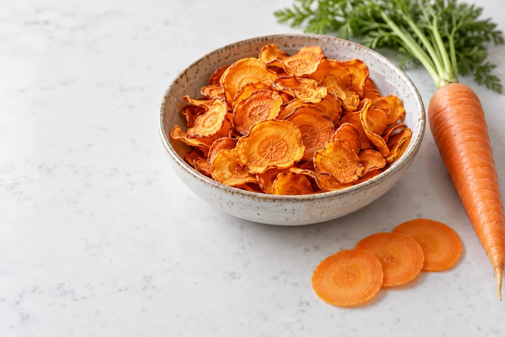 Thin crisp carrot chips in a bowl beside fresh carrot and raw slices