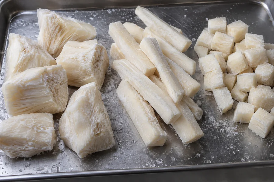 Three actual cassava formats: large peeled frozen chunks, uniform sticks, small cubes, clearly separated on a clean cold tray